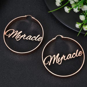 May include: A pair of gold hoop earrings with the word "Miracle" cut out in the metal. The earrings are large and have a simple design.