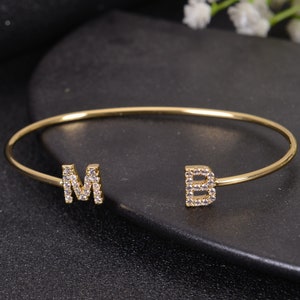 May include: A gold-tone open bangle bracelet featuring the letters "M" and "B", each embellished with small, clear crystals. The bracelet is displayed on a dark surface.