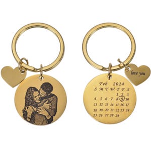 May include: Gold-tone keychain with a round photo charm and a round charm with the month of February 2024 calendar. The photo charm features a black and white photo of a couple embracing. The calendar charm has a heart symbol over the 8th. The keychain also includes a small heart charm with the text "love you".