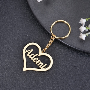 May include: Gold heart-shaped keychain with the name "Ademi" cut out in the center. The keychain is attached to a gold chain and a gold key ring.