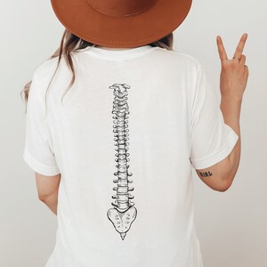 Może przedstawiać: Biała koszulka z czarno-białą ilustracją ludzkiego kręgosłupa. Projekt jest szczeg&oacute;łowy. Słowo "GIRL" jest wytatuowane na ramieniu.