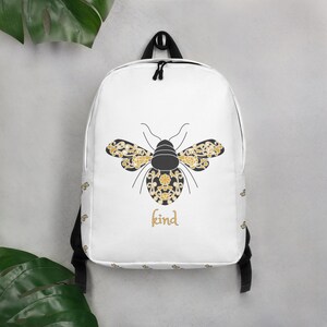 Puede incluir: Mochila blanca con un diseño de abeja negra y dorada con detalles florales. La palabra "kind" está escrita en letras doradas. La mochila tiene correas negras y un asa superior.