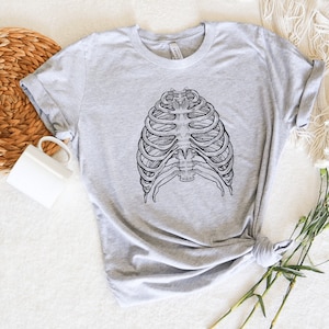 Może przedstawiać: Jasnoszara koszulka z szczeg&oacute;łową czarno-białą ilustracją ludzkiej klatki piersiowej. Koszulka ma klasyczny okrągły dekolt i podwinięte rękawy. Wykonana z miękkiego materiału.