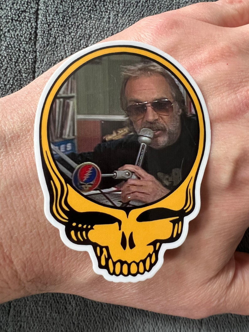 May include: A yellow sticker with a cartoon skull design. The skull has a black outline and white teeth. Inside the skull is a photo of a man with a microphone. The man is wearing sunglasses and a black jacket. The sticker has a yellow border.