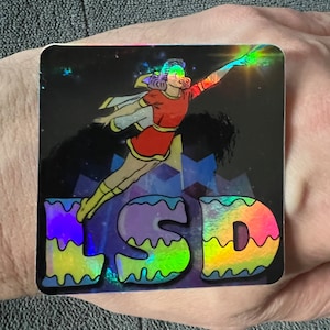 May include: A holographic sticker with a woman in a red and yellow costume flying with a glowing hand. The text "LSD" is in rainbow colors and has a wavy outline.