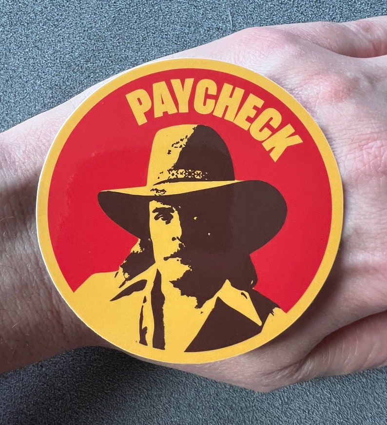 May include: A round sticker with a red background and a yellow border. The sticker features a cartoon cowboy wearing a brown hat and a yellow shirt. The text "PAYCHECK" is written in yellow letters around the cowboy's head.