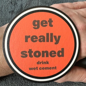 May include: A round orange coaster with a white border and black text that reads "get really stoned drink wet cement".