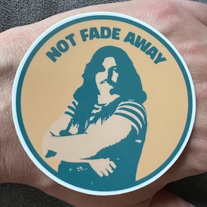 May include: A round sticker with a yellow background and a teal border. The sticker features a teal silhouette of a person with long hair and a striped shirt. The text "NOT FADE AWAY" is written in a teal arc around the person.