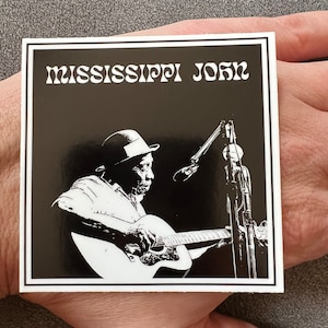 May include: Black and white sticker featuring a silhouette of Mississippi John Hurt playing a guitar with a microphone in the background. The text "MISSISSIPPI JOHN" is at the top of the sticker.