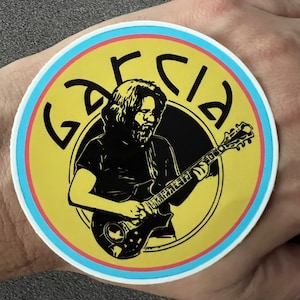 May include: A round sticker featuring a black and white illustration of a musician playing a guitar. The sticker has a yellow background with the name "GARCIA" in black, and is bordered by a light blue and pink ring.