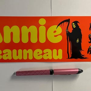 May include: An orange sticker with yellow text that reads "Annie Beauneau" and "From Saint Angel". A black Grim Reaper graphic is on the right. A pink pen is below the sticker.