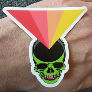 May include: A green skull with a black top and a rainbow triangle over the top of the skull.