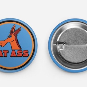 May include: A round button with a cartoon donkey head and the text "EAT ASS" in orange and black. The button has a light blue background with a black and orange border. The back of the button is silver.