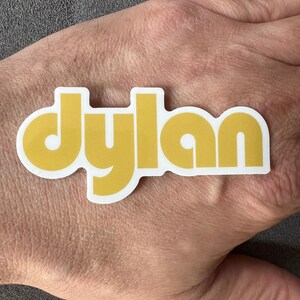 May include: A yellow sticker with the name "Dylan" in a bold, rounded font. The text is outlined in white, creating a contrast against the yellow. The sticker is likely for personalizing items.