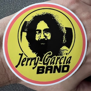 May include: A round sticker with a yellow background and a red and white border. The sticker features a black and white image of Jerry Garcia's face and the text "Jerry Garcia BAND" in black.