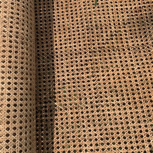 Pode incluir: Um rolo de tecido de vime natural, um material tecido usado para assentos de cadeiras e outros móveis estofados. O tecido é tecido apertado e tem uma cor castanho natural.