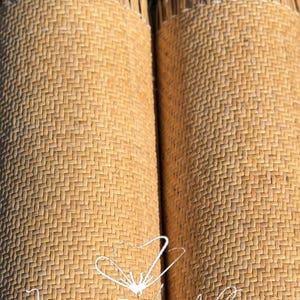 Puede incluir: Dos esteras enrolladas, tejidas con fibras naturales, probablemente ratán o material similar, en color beige claro. Las esteras tienen una superficie texturizada con un patrón repetitivo. El texto "Jassy Art Craft" está escrito en cursiva sobre las esteras.