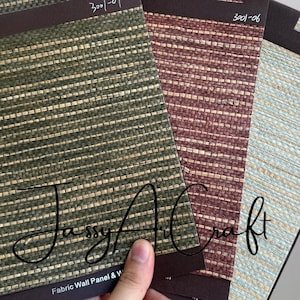 May include: A hand holds four fabric wall panel samples in shades of green, red, and blue. Each sample has a woven texture and is mounted on a dark brown backing. The samples are labeled with handwritten numbers.