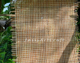 May include: A woven brown and white mesh fabric with a frayed edge. The fabric is hanging in front of a green leafy background.