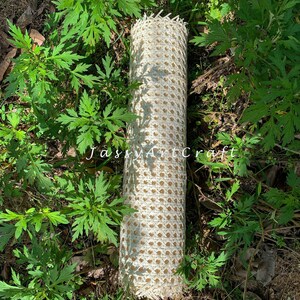 May include: A rolled-up, cream-colored woven cane material with a diamond pattern. The material is lying on the ground surrounded by green plants. The text "JassyArtCraft" is visible on the image.