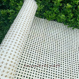 May include: A roll of natural-colored woven cane webbing, partially unrolled, against a backdrop of green foliage. The webbing features a repeating pattern of small, evenly spaced holes, ideal for furniture repair or craft projects. The text "JassyArtCraft" is visible.