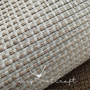 May include: Close-up of a woven textile with a repeating pattern of light beige and brown rectangular blocks. The texture appears to be made of natural fibers, possibly jute or a similar material. The textile is rolled up, revealing the underside.