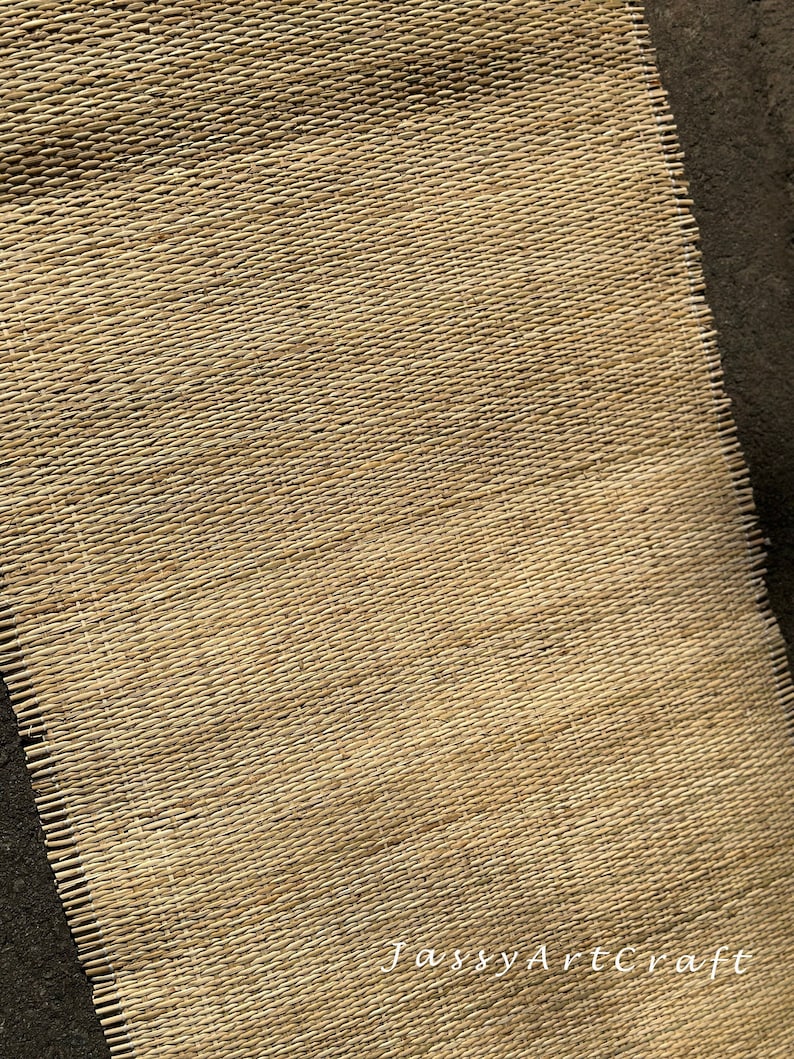 May include: A close-up of a woven natural fiber mat with a light brown color. The mat has a rough texture and is made of thin, tightly woven strands. The mat is slightly frayed at the edges. The text 'Jassy Artcraft' is visible in the bottom right corner of the mat.