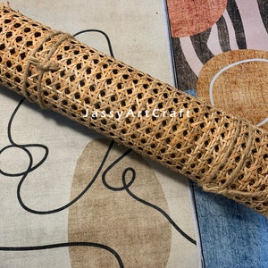 May include: A roll of light brown woven rattan cane webbing with a hexagonal pattern, secured with a thin jute twine. The roll is positioned over a fabric surface featuring a black line drawing of a face and abstract shapes with the text JassyArtCraft.