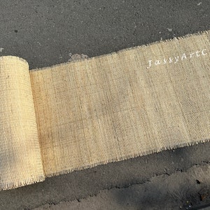 Pode incluir: Um tapete bege de fibra natural tecido, enrolado em uma superf&iacute;cie de asfalto cinza. O tapete tem uma borda desfiada e o texto "Jassy Art Craft" &eacute; vis&iacute;vel no tapete.