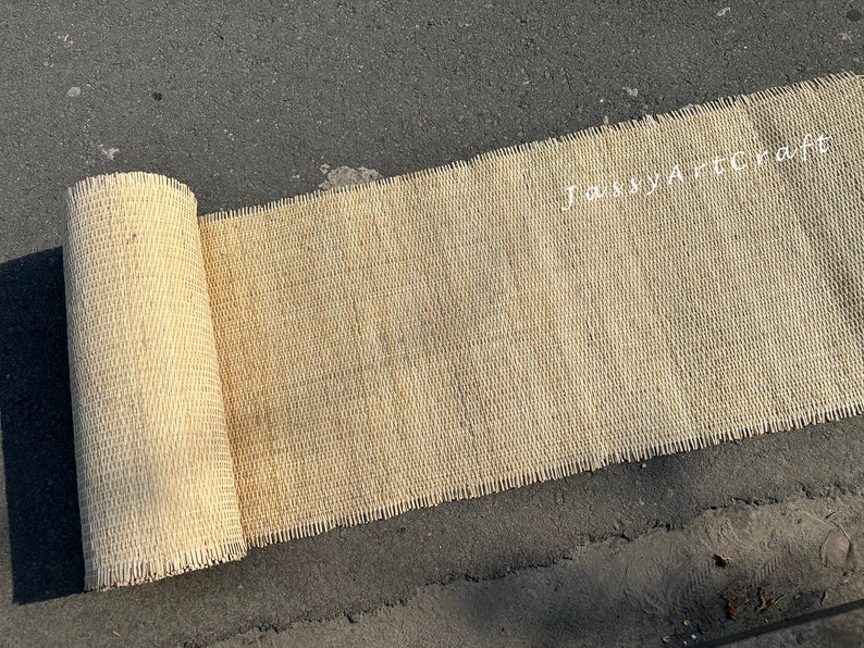 Pode incluir: Um tapete bege de fibra natural tecido, enrolado em uma superf&iacute;cie de asfalto cinza. O tapete tem uma borda desfiada e o texto "Jassy Art Craft" &eacute; vis&iacute;vel no tapete.