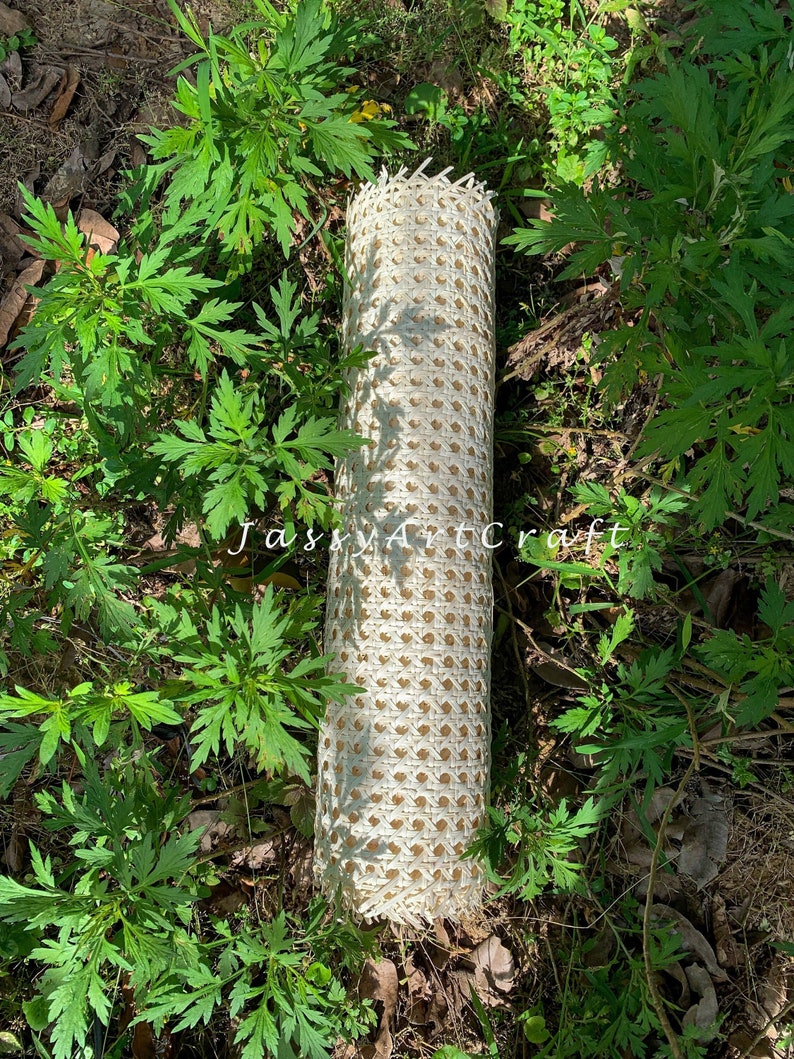May include: A rolled-up, cream-colored woven cane material with a diamond pattern. The material is lying on the ground surrounded by green plants. The text "JassyArtCraft" is visible on the image.