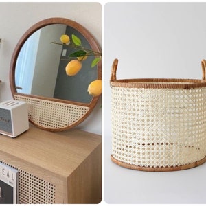 May include: A round woven wicker basket with two handles. The basket is light beige with a natural brown woven rim. The basket is sitting on a white surface.