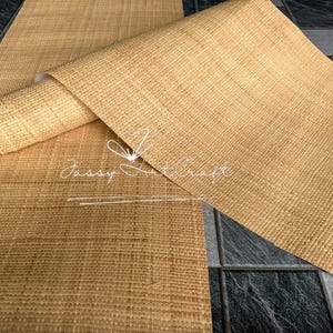 May include: A close-up of a natural-colored woven raffia material, partially rolled. The material has a textured surface and is labeled "Jassy Art Craft" in white script. The background is a dark gray tile.