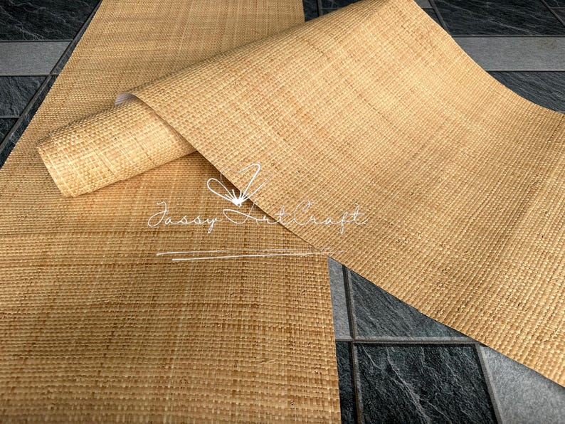 May include: A close-up of a natural-colored woven raffia material, partially rolled. The material has a textured surface and is labeled "Jassy Art Craft" in white script. The background is a dark gray tile.