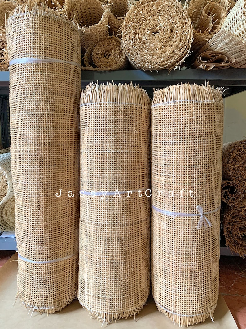 May include: Three rolls of natural woven cane webbing, a type of material used for chair seats and other furniture. The rolls are stacked on top of each other on a shelf. The webbing is a light brown color and has a woven pattern. The rolls are tied with white string.