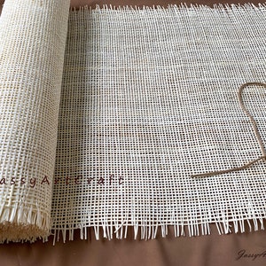 Puede incluir: Un rollo de tejido de ca&ntilde;a blanco tejido con bordes deshilachados. El tejido est&aacute; enrollado y atado con una cuerda. El texto "Jassy Artcraft" est&aacute; impreso en el tejido.