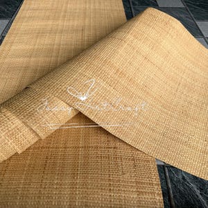 May include: A rolled-up, woven, natural-colored mat with a textured surface. The mat is partially unrolled, revealing its full width. The background is a dark, tiled surface. The mat has the text "Jassy Artcraft" in a cursive font.