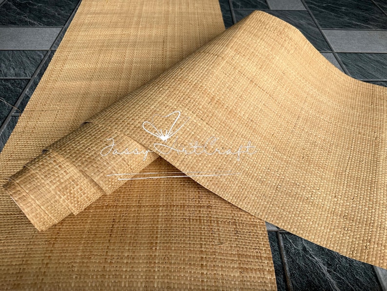 May include: A rolled-up, woven, natural-colored mat with a textured surface. The mat is partially unrolled, revealing its full width. The background is a dark, tiled surface. The mat has the text "Jassy Artcraft" in a cursive font.