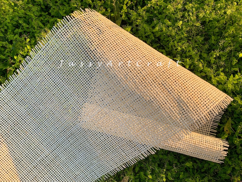 May include: A close-up of a beige cane webbing material with a woven pattern. The material is folded in half and has frayed edges. The material is lying on a bed of green grass.