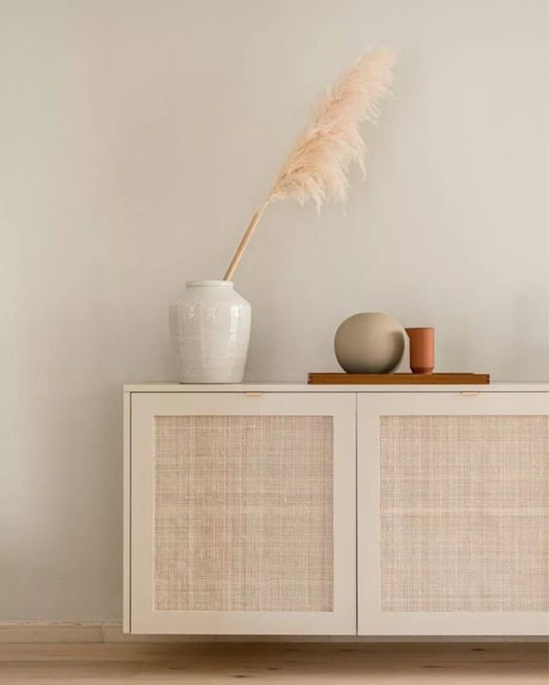 Puede incluir: Un armario blanco con dos puertas, cada una con un panel de rat&aacute;n tejido. El armario est&aacute; sobre un suelo de madera. Un jarr&oacute;n blanco con una pluma de hierba de la pampa seca se coloca encima del armario, junto con una esfera de cer&aacute;mica marr&oacute;n y un peque&ntilde;o jarr&oacute;n de terracota.