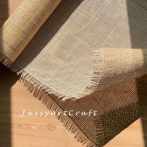 May include: A close-up of a beige woven cane mesh fabric with a fringed edge. The fabric is rolled up and the edges are visible. The text "JassyArtcraft" is visible in the lower left corner of the image.