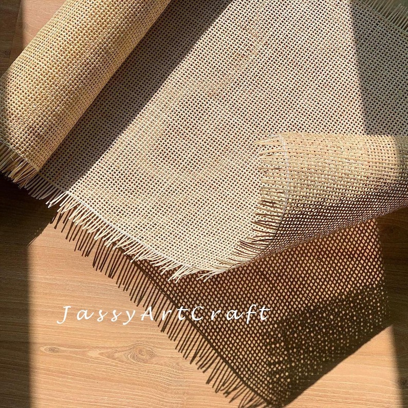 May include: A close-up of a beige woven cane mesh fabric with a fringed edge. The fabric is rolled up and the edges are visible. The text "JassyArtcraft" is visible in the lower left corner of the image.