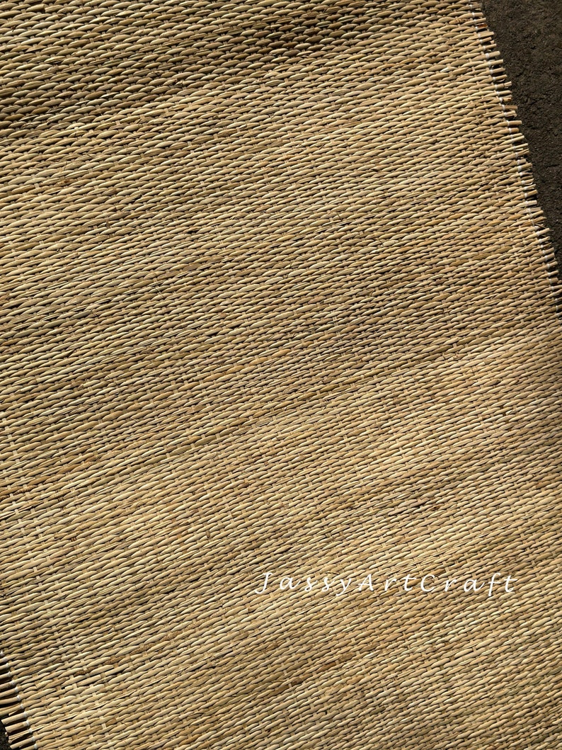 Pode incluir: Um close-up de um tapete tecido de fibra natural com um padr&atilde;o diagonal. O tapete &eacute; de cor castanho claro e tem uma textura &aacute;spera. O texto "Jassy Artcraft" &eacute; vis&iacute;vel no canto inferior direito da imagem.