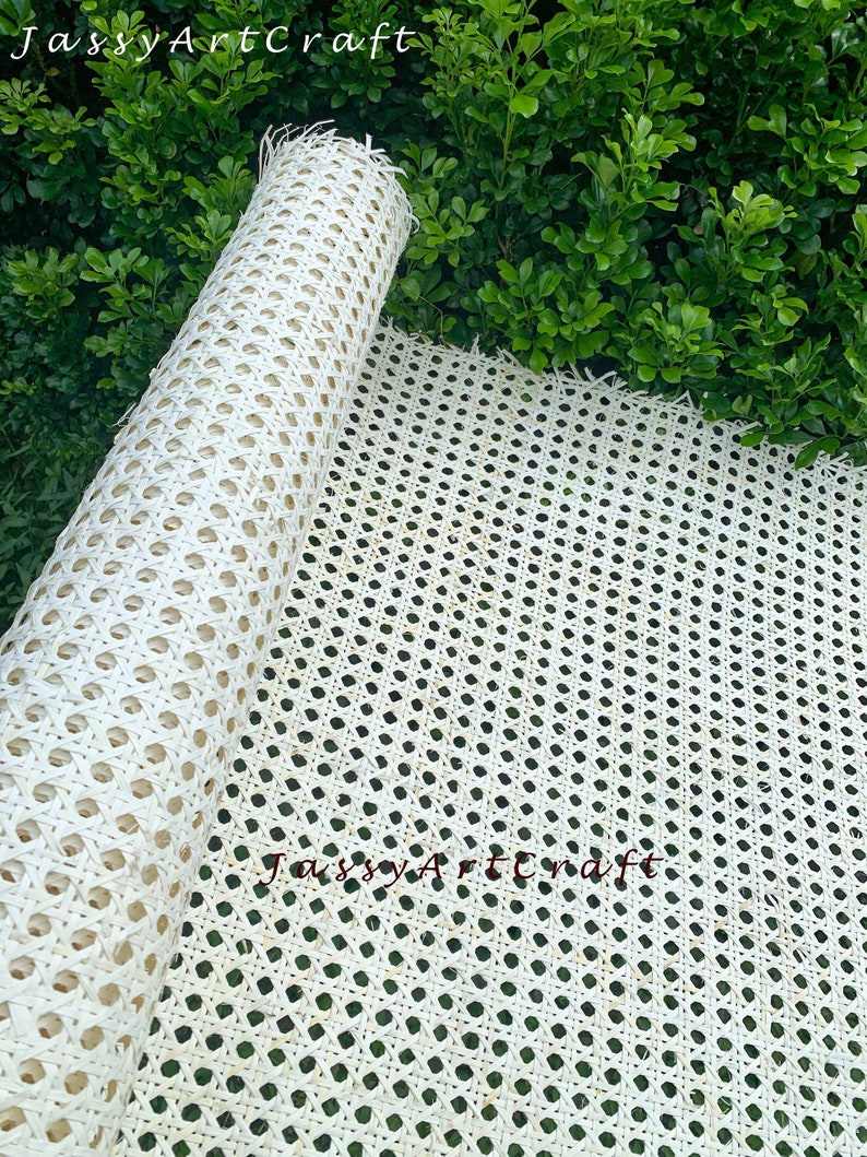 May include: A roll of natural-colored woven cane webbing, partially unrolled, against a backdrop of green foliage. The webbing features a repeating pattern of small, evenly spaced holes, ideal for furniture repair or craft projects. The text "JassyArtCraft" is visible.