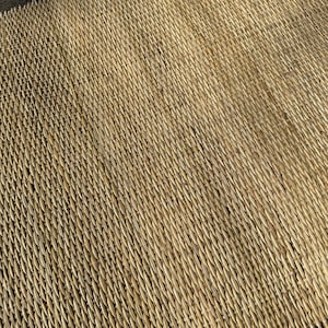 Pode incluir: Um close-up de um tapete natural tecido de palha ou juncos. O tapete tem uma cor castanho claro e uma superf&iacute;cie texturizada. O tapete &eacute; tecido em um padr&atilde;o apertado, criando uma superf&iacute;cie lisa e uniforme.