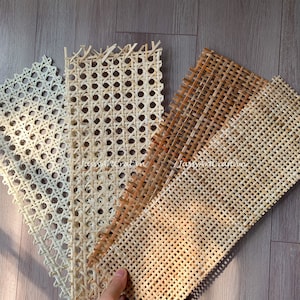 May include: A hand holds several samples of woven cane webbing in varying shades of beige and brown. The samples showcase different patterns and textures, ideal for furniture repair or craft projects. The background is a wooden surface.