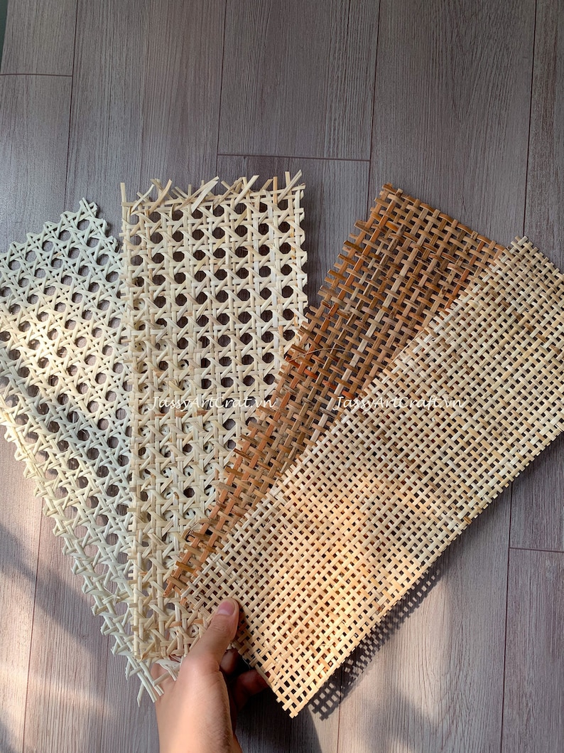 May include: A hand holds several samples of woven cane webbing in varying shades of beige and brown. The samples showcase different patterns and textures, ideal for furniture repair or craft projects. The background is a wooden surface.
