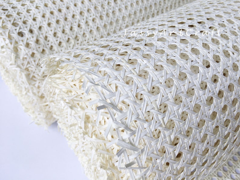 May include: Close-up of a rolled-up, off-white woven material. The material has a repeating pattern of interwoven strips, creating a grid-like texture with open spaces. The texture is visible, and the material appears to be flexible.