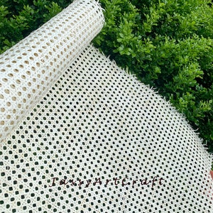 May include: A roll of off-white woven cane webbing, partially unrolled, displaying a repeating pattern of small, evenly spaced holes. The material is set against a backdrop of lush green foliage, suggesting outdoor use or a natural aesthetic.