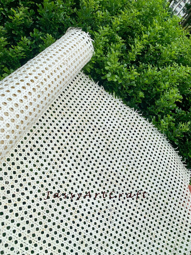 May include: A roll of off-white woven cane webbing, partially unrolled, displaying a repeating pattern of small, evenly spaced holes. The material is set against a backdrop of lush green foliage, suggesting outdoor use or a natural aesthetic.